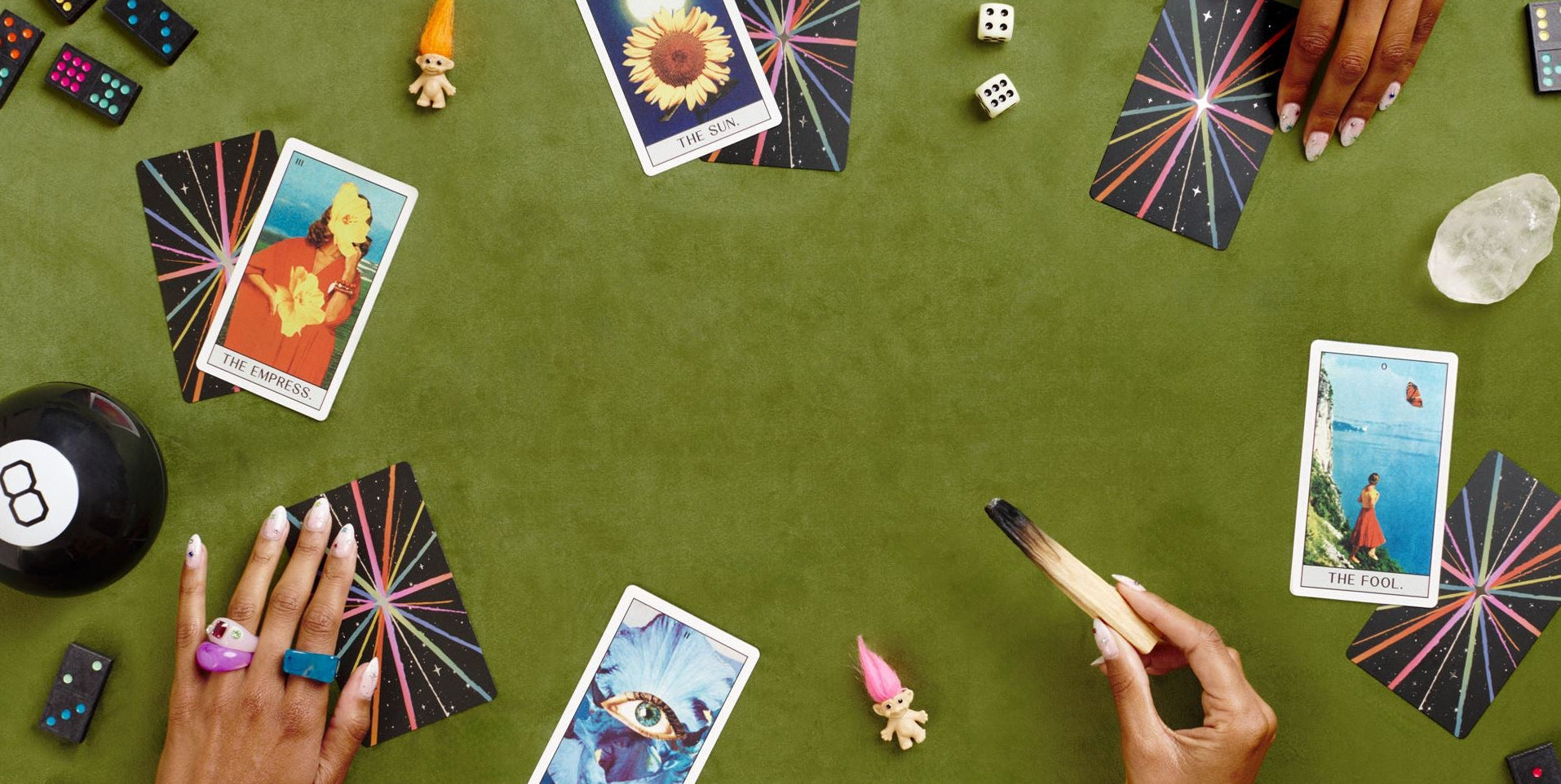 Tarot cards, trolls dolls, magic 8 ball, dominoes and dice arranged on a green surface with hands, crystals and palo santo.