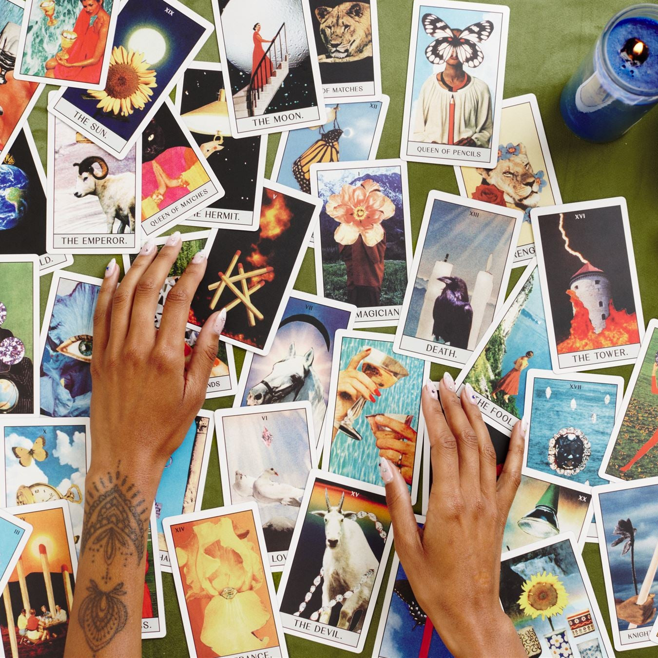 Tarot cards chaotically spread on table with hands reaching for the cards, blue candle, quartz crystal, magic 8 ball and dice sit to the side of the cards on a green surface 