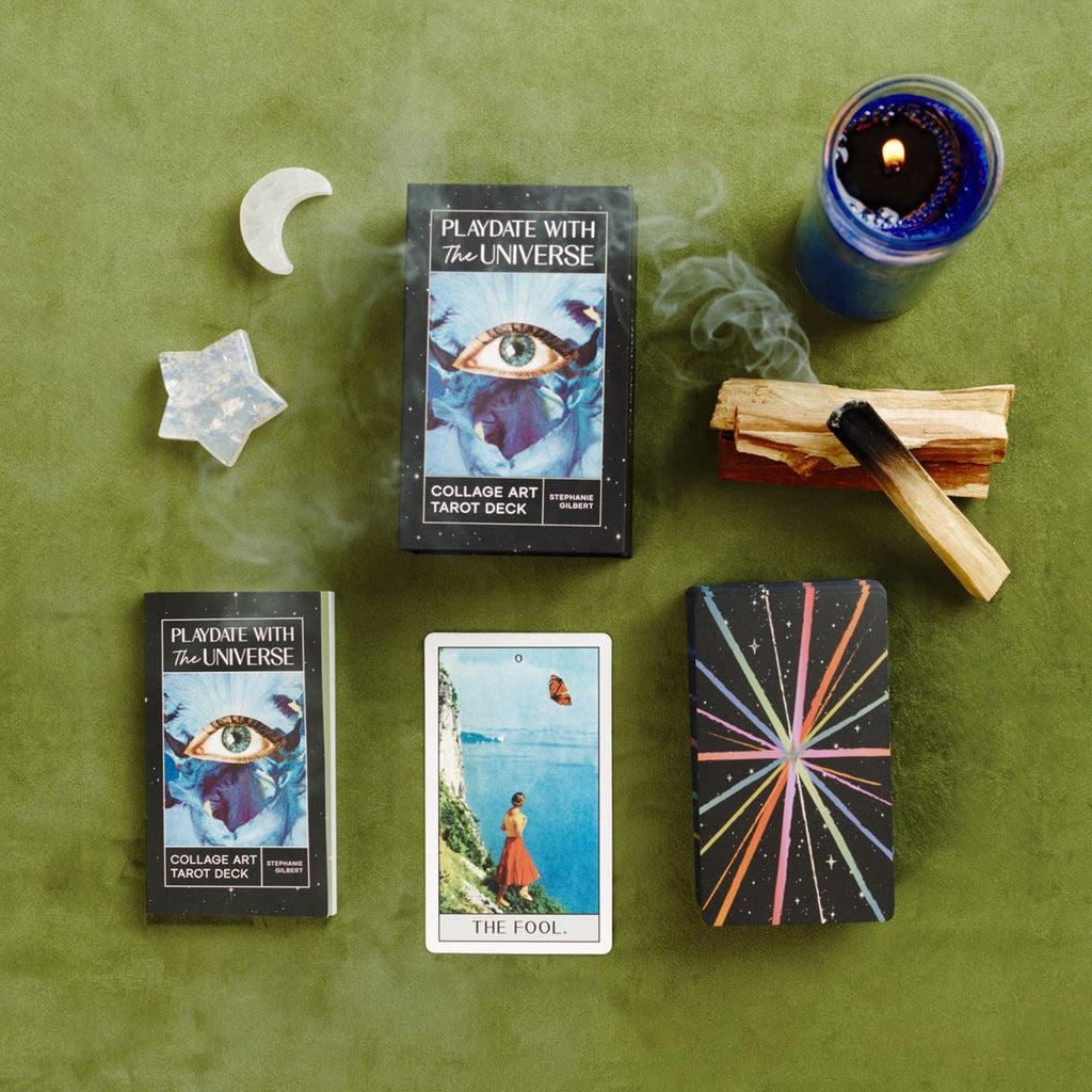 Tarot deck box, guidebook and cards, a moon and star crystals, a lit candle, and smoking palo santo arranged in a flat lay on a green surface