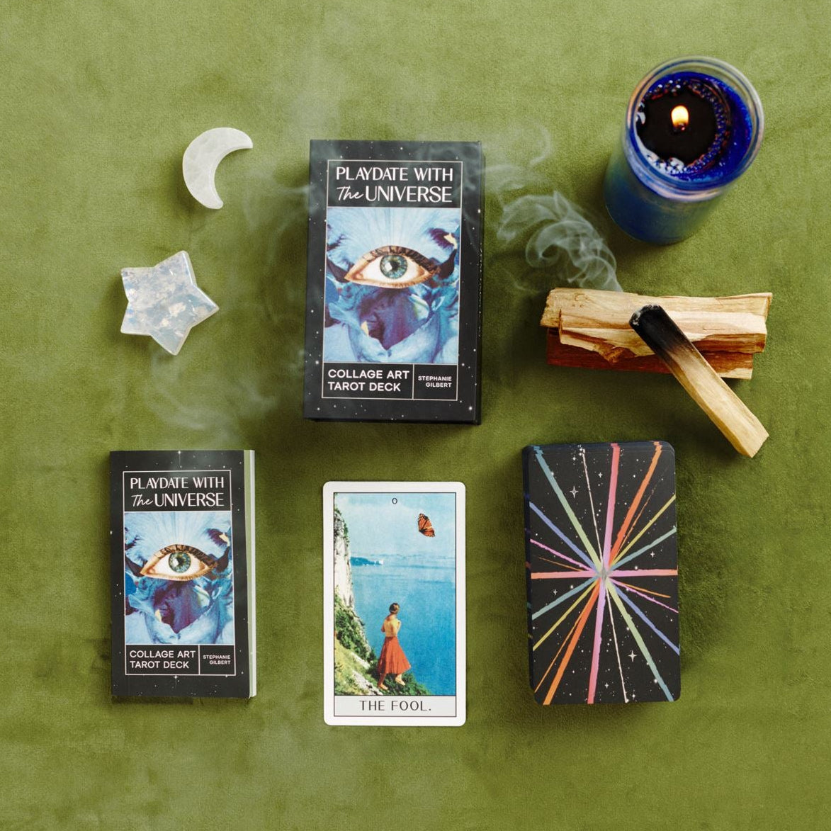 Tarot deck box, guidebook and cards, a moon and star crystals, a lit candle, and smoking palo santo arranged in a flat lay on a green surface
