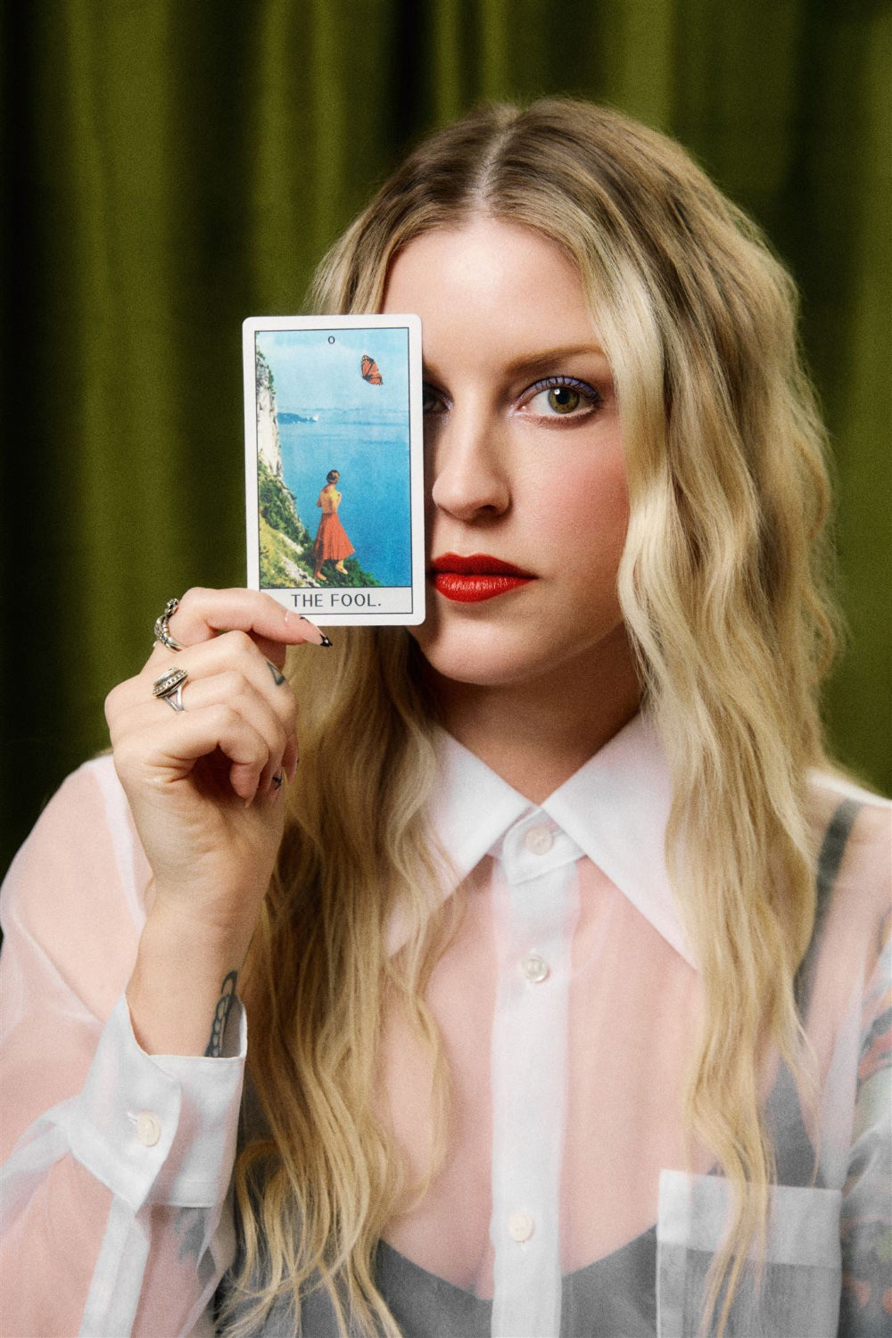 woman in red lipstick holding the fool tarot card over one eye with green backdrop